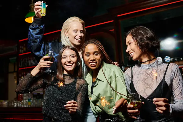 Happy and trendy multiethnic girlfriends toasting with cocktails and holding shiny sparklers in bar — Stock Photo