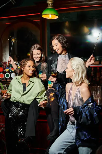 Copines multiraciales glamour et heureux avec des cocktails et des étincelles festives dans le bar, temps de fête — Photo de stock