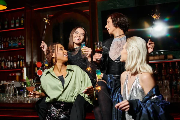 Fashionable and happy multiracial girlfriends with cocktails and shiny sparklers in bar, party time — Stock Photo