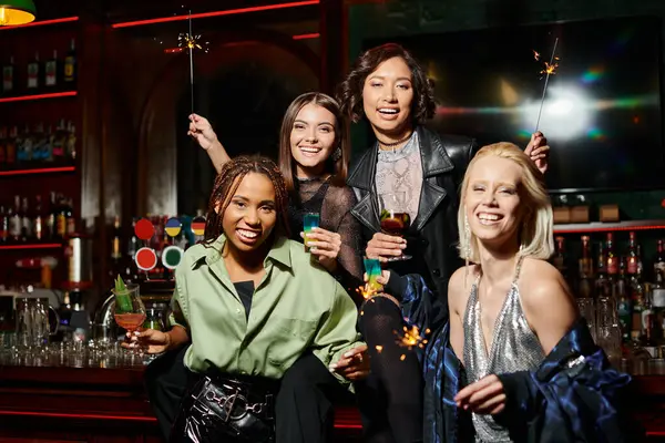 Веселые стильные многорасовые подружки с коктейлями и блестящими сверкающими блестками, смотрящими в камеру в баре — стоковое фото