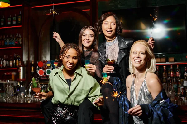 Cheerful stylish multiracial girlfriends with cocktails and shiny sparklers looking at camera in bar — Stock Photo