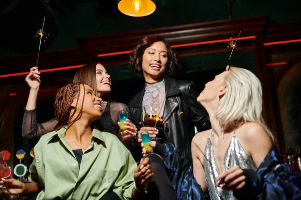 Sorridenti ed eleganti fidanzate multirazziali con cocktail e scintillanti scintille nel bar, festa festiva — Foto stock