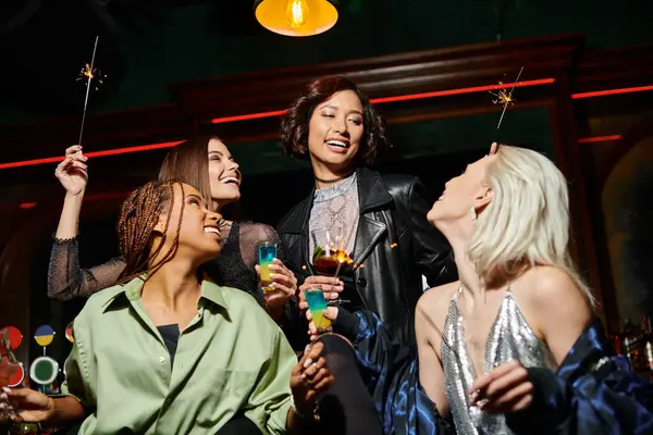Smiling and stylish multiracial girlfriends with cocktails and shiny sparklers in bar, festive party — Stock Photo