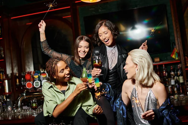 Aufgeregte multiethnische Freundinnen mit glänzenden Wunderkerzen, die in der Bar Cocktailgläser klirren, lebhafte Party — Stockfoto