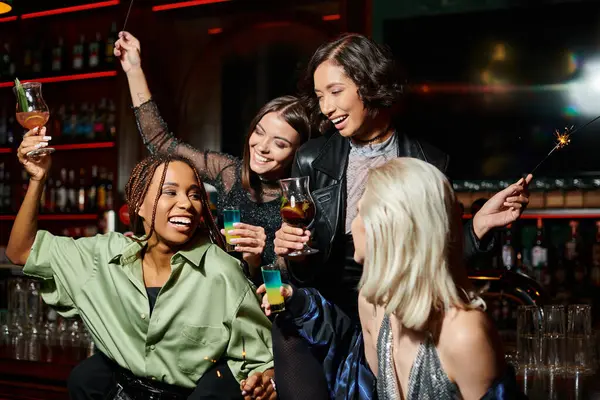 Happy multicultural girlfriends with shiny sparklers and cocktail glasses in bar, festive atmosphere — Stock Photo