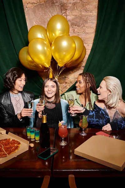 Cheerful multiracial fashionistas with champagne near cocktails and golden balloons in bar, birthday — Stock Photo