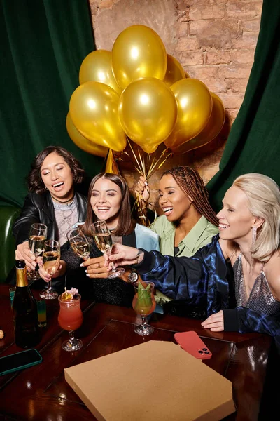 Novias multiétnicas tintineo copas de champán cerca de globos de oro en el bar, fiesta de cumpleaños - foto de stock