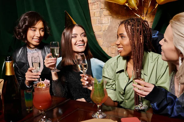 Young birthday girl in party hat holding champagne near multiethnic girlfriends and cocktails in bar — Stock Photo