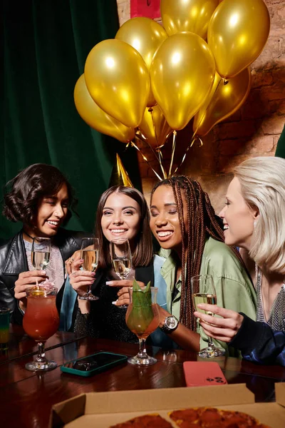 Felice donna in cappello partito con champagne vicino a fidanzate multietniche e palloncini di compleanno nel bar — Foto stock