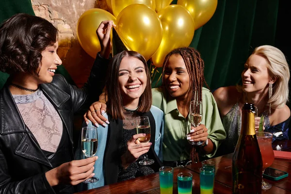 Joyous multiethnic girlfriends with champagne celebrating birthday near golden balloons in bar — Stock Photo