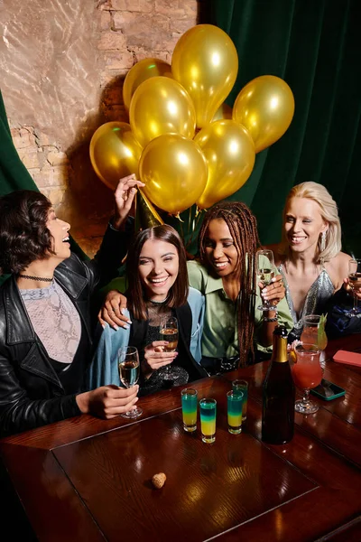 Happy stylish multiethnic women with champagne near golden balloons and cocktails in bar, birthday — Stock Photo