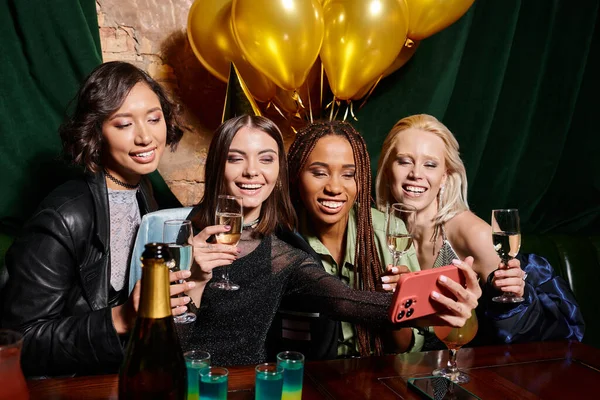 Cheerful multicultural girlfriends with champagne taking selfie on smartphone during birthday party — Stock Photo