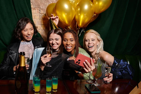 Excited multicultural girlfriends with champagne taking selfie on smartphone in bar, birthday party — Stock Photo