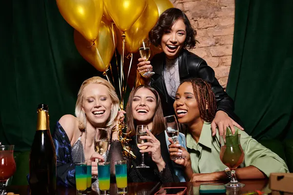 Joyful stylish multiethnic girlfriends near cocktails and golden festive balloons in bar, birthday — Stock Photo