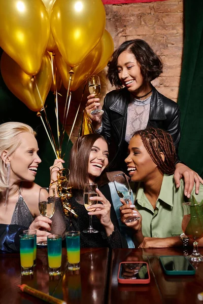 Novias multiétnicas de moda con champán sonriendo cerca de cócteles y globos de oro, cumpleaños - foto de stock