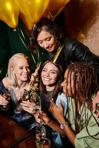 Pleased woman in party hat with champagne smiling with closed eyes near multiethnic girlfriends — Stock Photo
