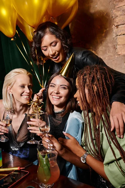 Joyful woman in party hat holding champagne near multiethnic girlfriends and balloons in bar — Stock Photo
