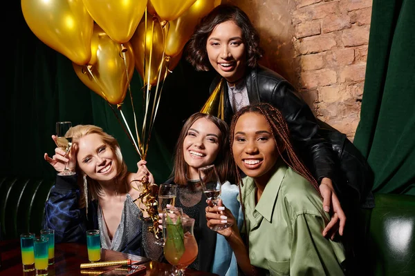 Gioiosa donne multirazziali con champagne guardando la fotocamera vicino palloncini d'oro nel bar, compleanno — Foto stock