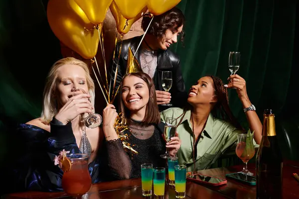 Возбужденные многорасовые девушки пьют шампанское возле золотых шариков в баре, день рождения — стоковое фото