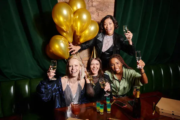Cheerful multicultural girlfriends with champagne looking at camera and celebrating birthday in bar — Stock Photo