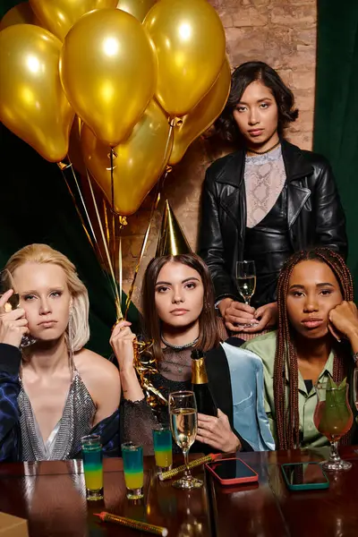 Серьезные многонациональные девушки с шампанским, смотрящие в камеру рядом с золотыми воздушными шарами, день рождения — стоковое фото