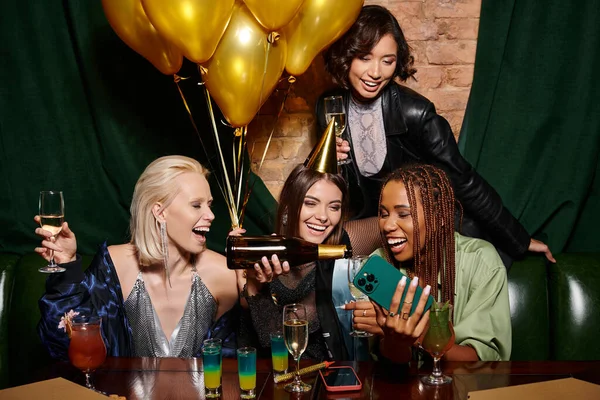 Mulher alegre derramando champanhe perto de namoradas multiculturais durante a festa de aniversário no bar — Fotografia de Stock