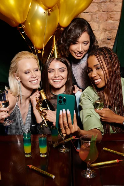 Happy woman in party hat taking selfie with stylish multiracial girlfriends, birthday party in bar — Stock Photo