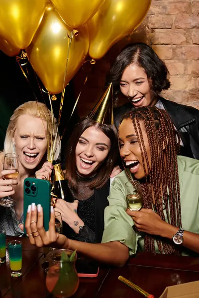 Mujer despreocupada en sombrero de fiesta tomando selfie con las novias multirraciales de moda, fiesta de cumpleaños en el bar - foto de stock