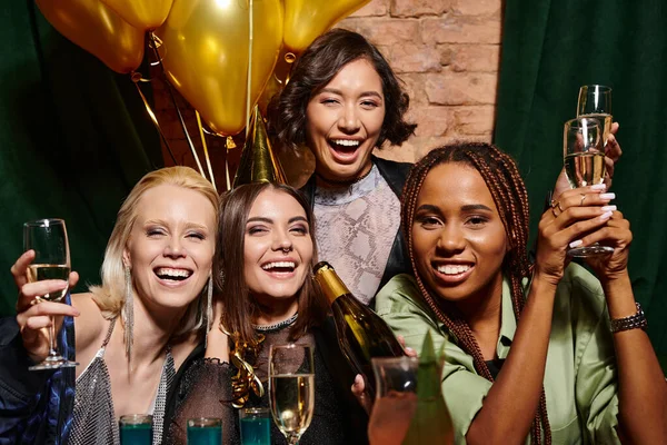 Joyful multicultural women with champagne and golden balloons looking at camera in bar, birthday — Stock Photo