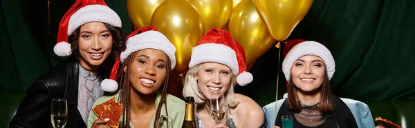 Excited multicultural female friends in party hats looking at camera near golden balloons, banner — Stock Photo
