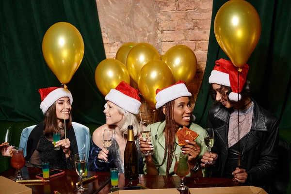 Cheerful multiracial women in Christmas hats near golden balloons and drinks with pizza in bar — Stock Photo