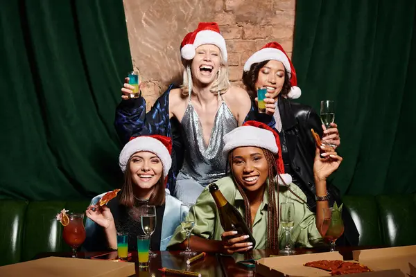 Mujeres multirraciales en sombreros de Navidad celebración de champán y mirando a la cámara en el bar, fiesta festiva - foto de stock