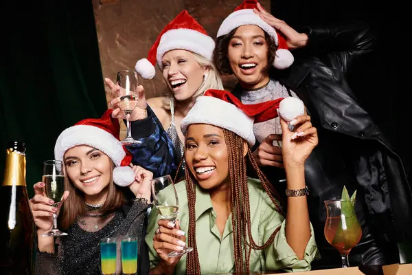 Retrato de grupo de novias multiétnicas emocionadas en sombrero de Navidad con copas de champán en el bar - foto de stock