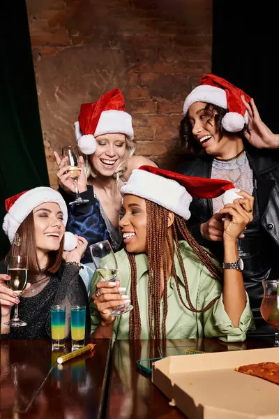 Novias multiétnicas jóvenes y despreocupadas en sombreros de Navidad con champán divertirse en el bar, fiesta - foto de stock