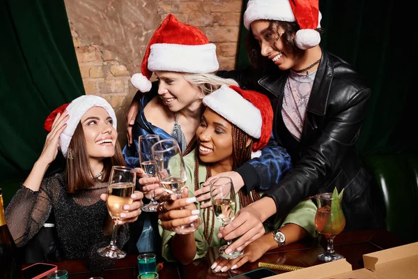 Feliz multiétnico novias en Navidad sombreros tintineo copas de champán en fiesta festiva en el bar - foto de stock