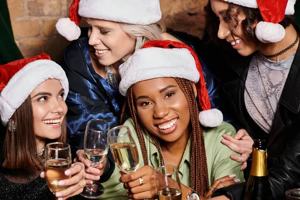 Mujer afroamericana en sombrero de Navidad con champán sonriendo a la cámara cerca de amigos multiétnicos - foto de stock