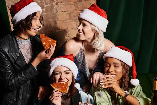 Joyeuses petites amies multiethniques dans des chapeaux de Noël boire du champagne et manger de la pizza dans un bar, fête — Photo de stock