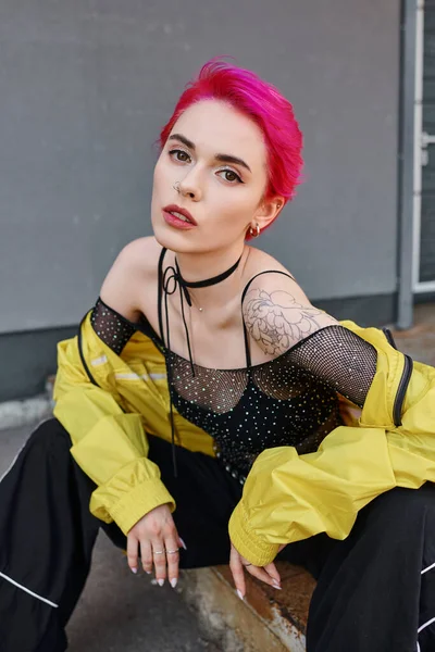 Stylish young tattooed woman in trendy outfit sitting and looking at camera, fashion concept — Stock Photo