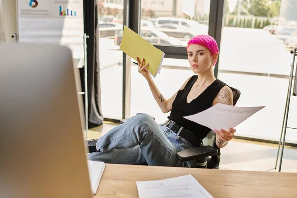 Nachdenkliche rosa haarige Geschäftsfrau in lässigem Outfit am Computer sitzend mit ihren Notizen in der Hand — Stockfoto