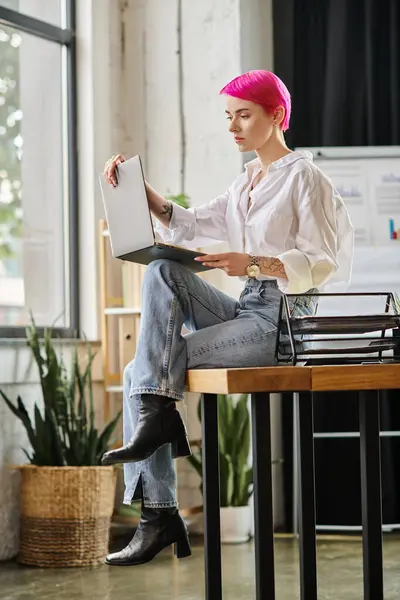 Молодая розововолосая деловая женщина в повседневной городской одежде сидит и смотрит на свой ноутбук в офисе — стоковое фото
