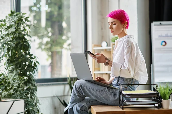 Junge schöne Frau in weißem Hemd mit rosafarbenen Haaren schaut auf Telefon mit Laptop auf Knien, Geschäft — Stockfoto
