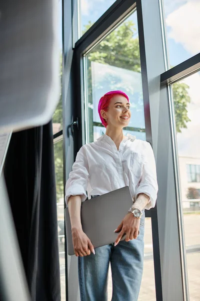 Joyeuse jeune femme d'affaires avec les cheveux courts roses tenant ordinateur portable et regardant loin de son bureau — Photo de stock