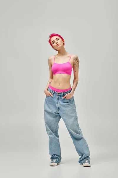 Привлекательная женщина с короткими розовыми волосами и татуировками в розовом топе и джинсах с руками в карманах — стоковое фото