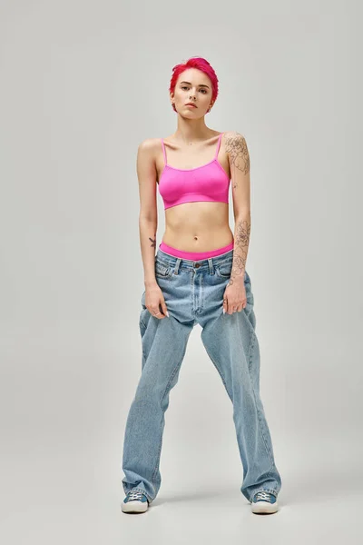 Jovem mulher de cabelos cor-de-rosa em top de cultura elegante e jeans azul posando em pano de fundo cinza, conceito de moda — Fotografia de Stock