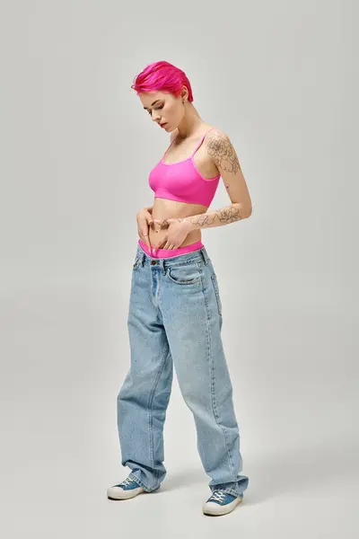 Joven mujer de pelo rosa en top de cosecha rosa y pantalones vaqueros azules posando con las manos en el vientre, concepto de moda - foto de stock