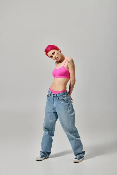 Attraktive junge Frau in rosa Crop Top und blauen Jeans, die vor grauem Hintergrund in die Kamera schaut, Mode — Stockfoto