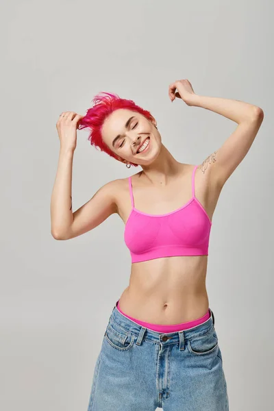 Mujer de pelo rosa feliz con tatuajes posando con los ojos cerrados sobre fondo gris, concepto de moda - foto de stock