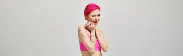 Збуджена молода жінка з рожевим коротким волоссям позує у верхній частині культури і підморгує на сірому тлі, банер — стокове фото