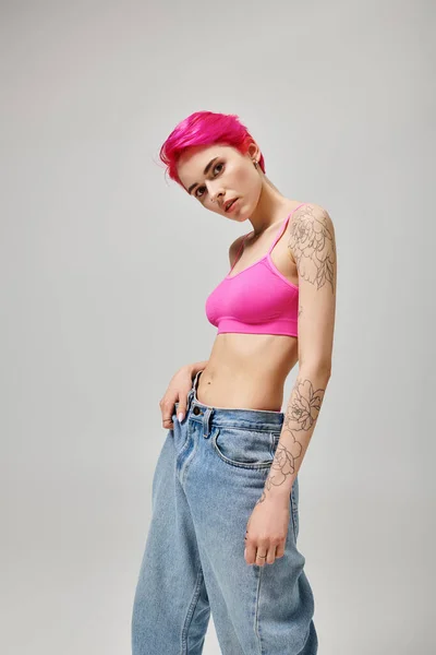 Giovane donna sicura di sé con capelli corti rosa in posa in crop top e jeans su sfondo grigio — Foto stock