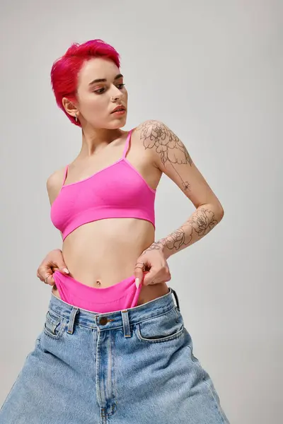 Jovem tatuada com cabelo rosa posando no topo da colheita e puxando calcinha de jeans em cinza — Fotografia de Stock
